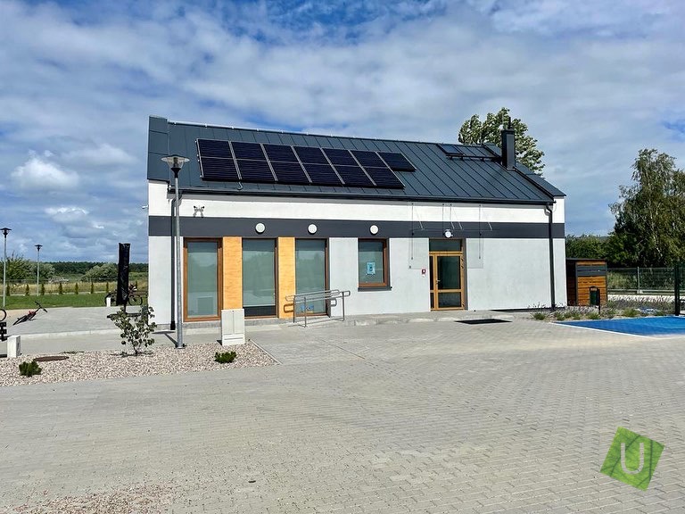 Nowoczesny budynek parterowy z panelami słonecznymi na dachu, szarą elewacją z białymi akcentami i rampą dla niepełnosprawnych, otoczony kostką brukową.