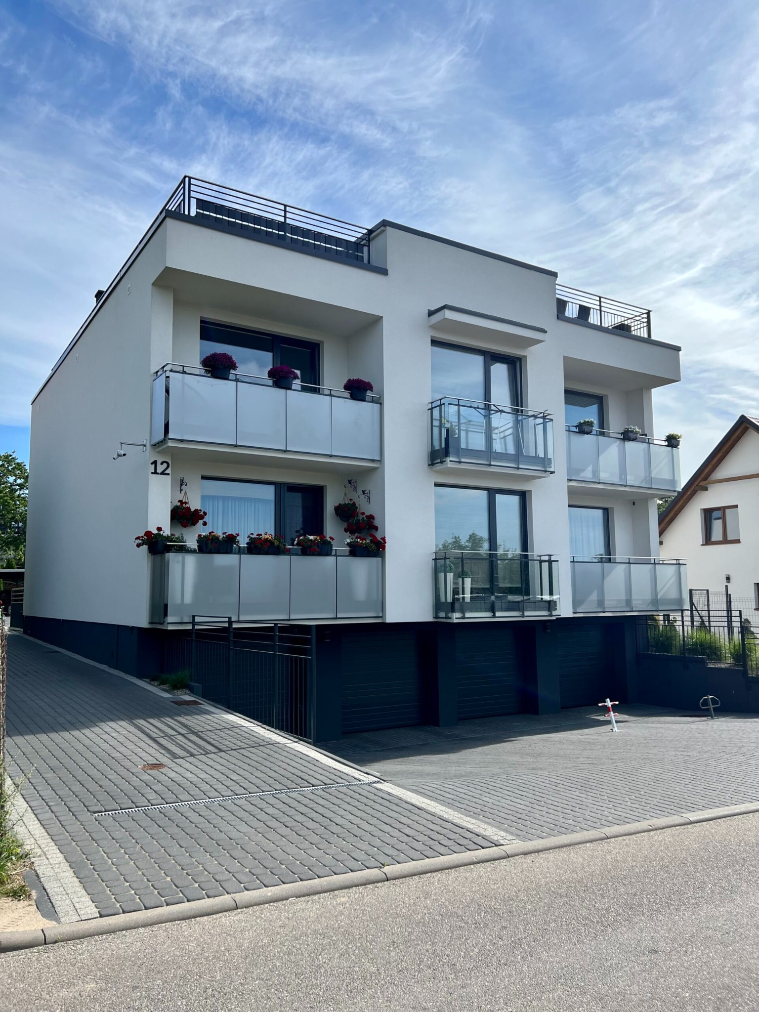 Nowoczesny, trzypiętrowy budynek mieszkalny z białymi ścianami, szklanymi balkonami z kwiatami i garażem w przyziemiu, numer 12 na ścianie, kostka brukowa na podjeździe.