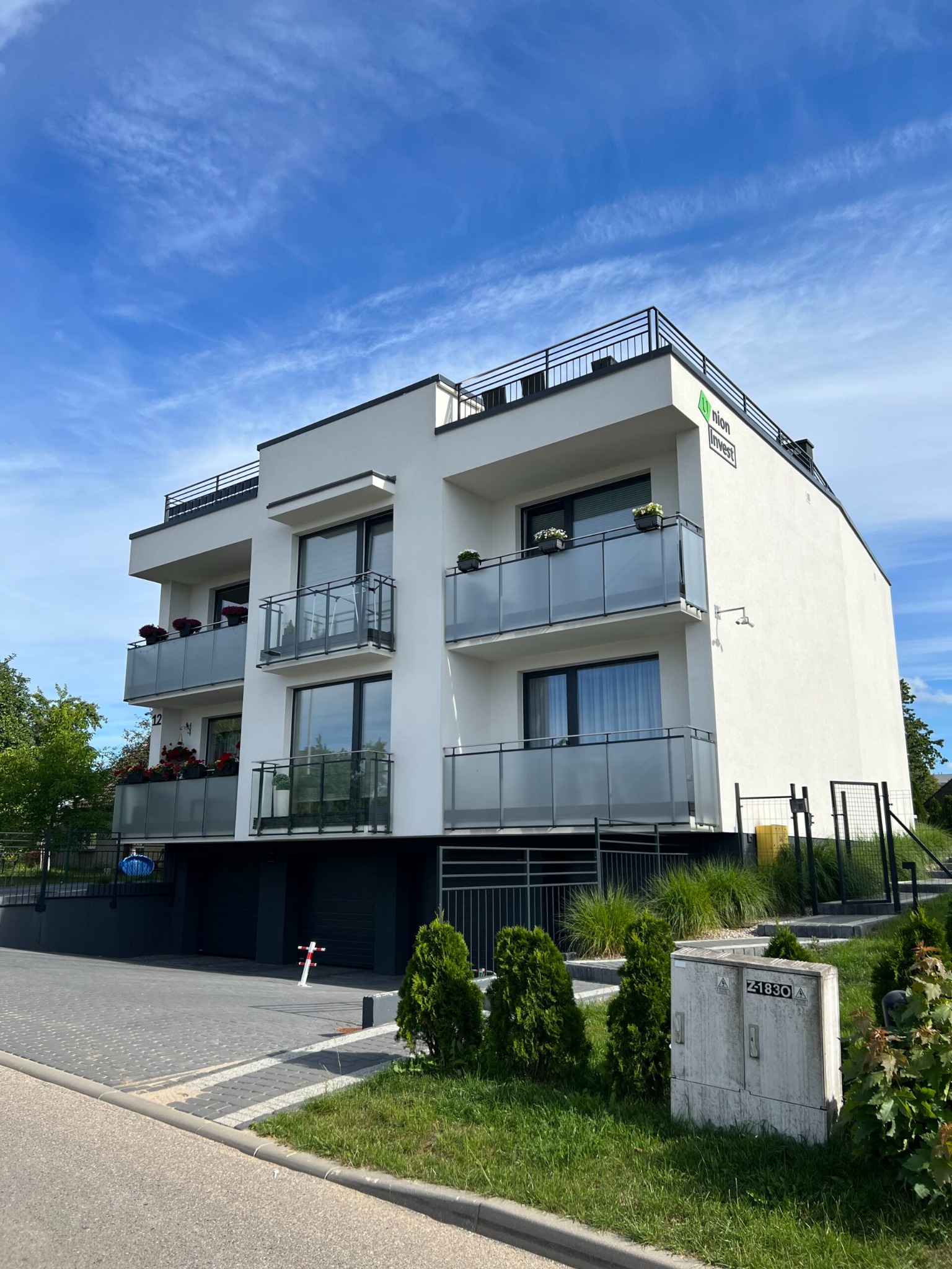 Nowoczesny, trzypiętrowy budynek apartamentowy z białymi ścianami, szklanymi balkonami z kwiatami, czarnymi ramami okien i balustradami, widoczny z poziomu chodnika z kostki brukowej w słoneczny...