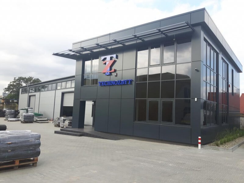 Nowoczesny budynek biurowo-magazynowy z szarą elewacją panelową, dużymi oknami i logo firmy Technozbyt nad wejściem, otoczony kostką brukową i składowanymi materiałami budowlanymi.