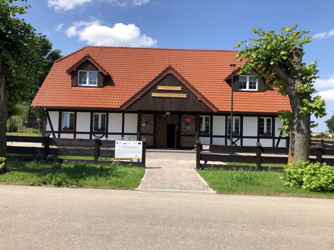 Budynek w stylu szachulcowym z czerwoną dachówką, widoczny z zewnątrz, otoczony zielenią i drewnianym ogrodzeniem. Na pierwszym planie chodnik z kostki i tablica informacyjna.