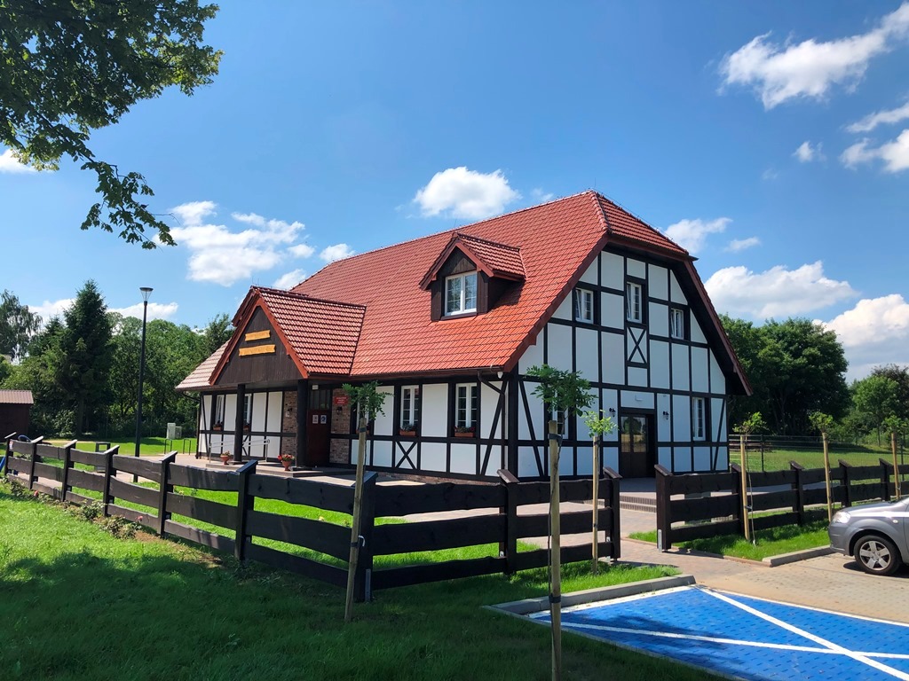 Dwukondygnacyjny budynek z czerwonym dachem i białymi ścianami z czarnymi belkami, otoczony drewnianym płotem i zielonym trawnikiem w słoneczny dzień.