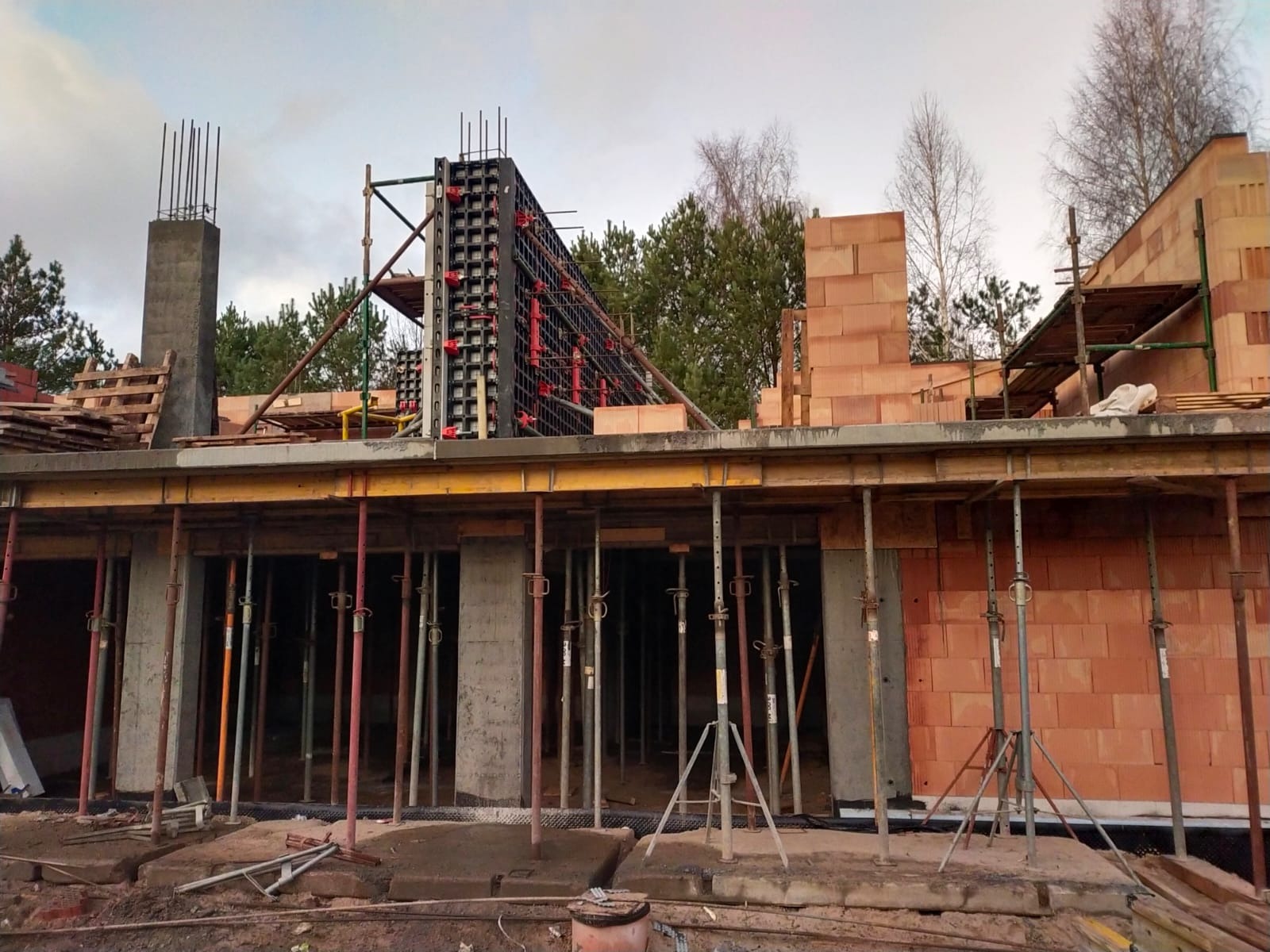 Budowa domu w stanie surowym otwartym: widoczne ściany z pustaków ceramicznych, szalunki zbrojeniowe, podpory stropowe i elementy drewniane konstrukcji dachu na tle drzew i pochmurnego nieba.