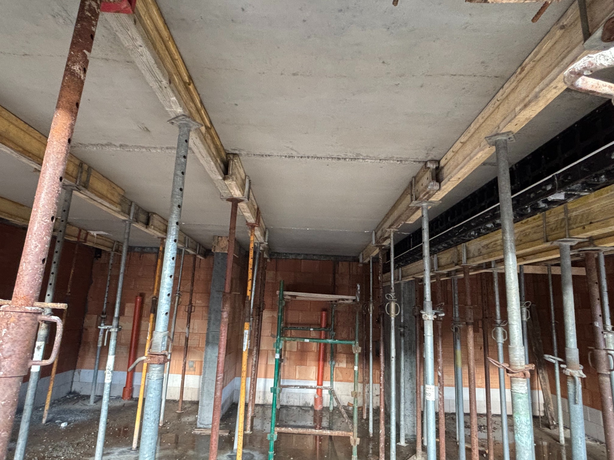 Wnętrze w trakcie budowy z widocznym stropem podpartym metalowymi stemplami i drewnianymi belkami, ceglane ściany w tle, na podłodze widoczne gruz i woda.