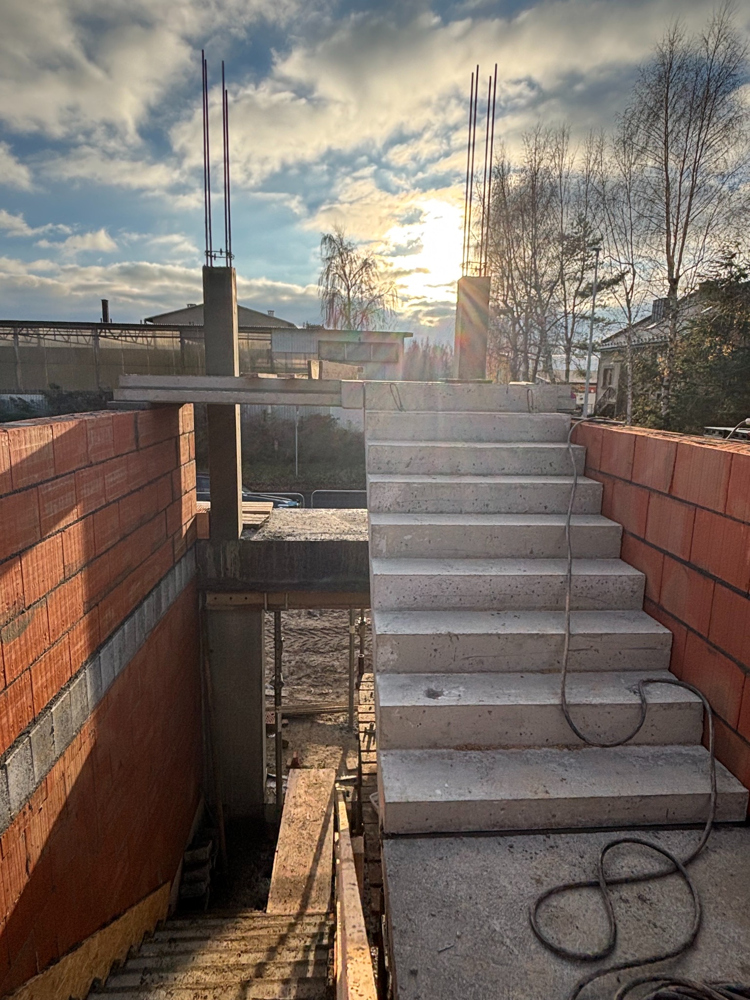 Surowe betonowe schody prowadzące do podpiwniczenia w trakcie budowy domu z cegły, z wystającymi prętami zbrojeniowymi w słupach, widok na zewnątrz na częściowo pochmurne niebo i drzewa.