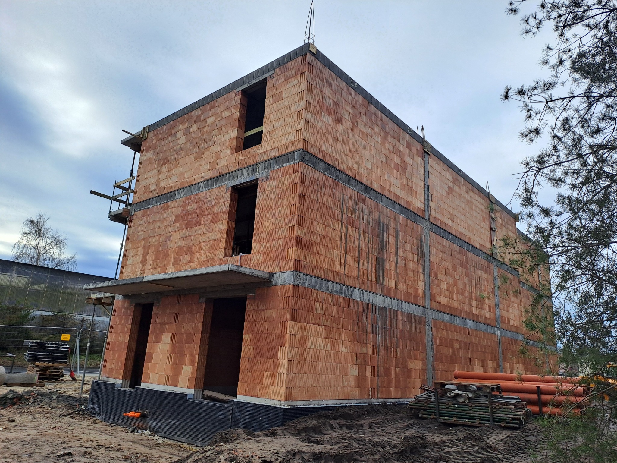 Trzypiętrowy budynek w trakcie budowy z czerwonej cegły ceramicznej, widoczne betonowe stropy, rusztowania i stos rur na ziemi, inwestycja UTBS Ustka.