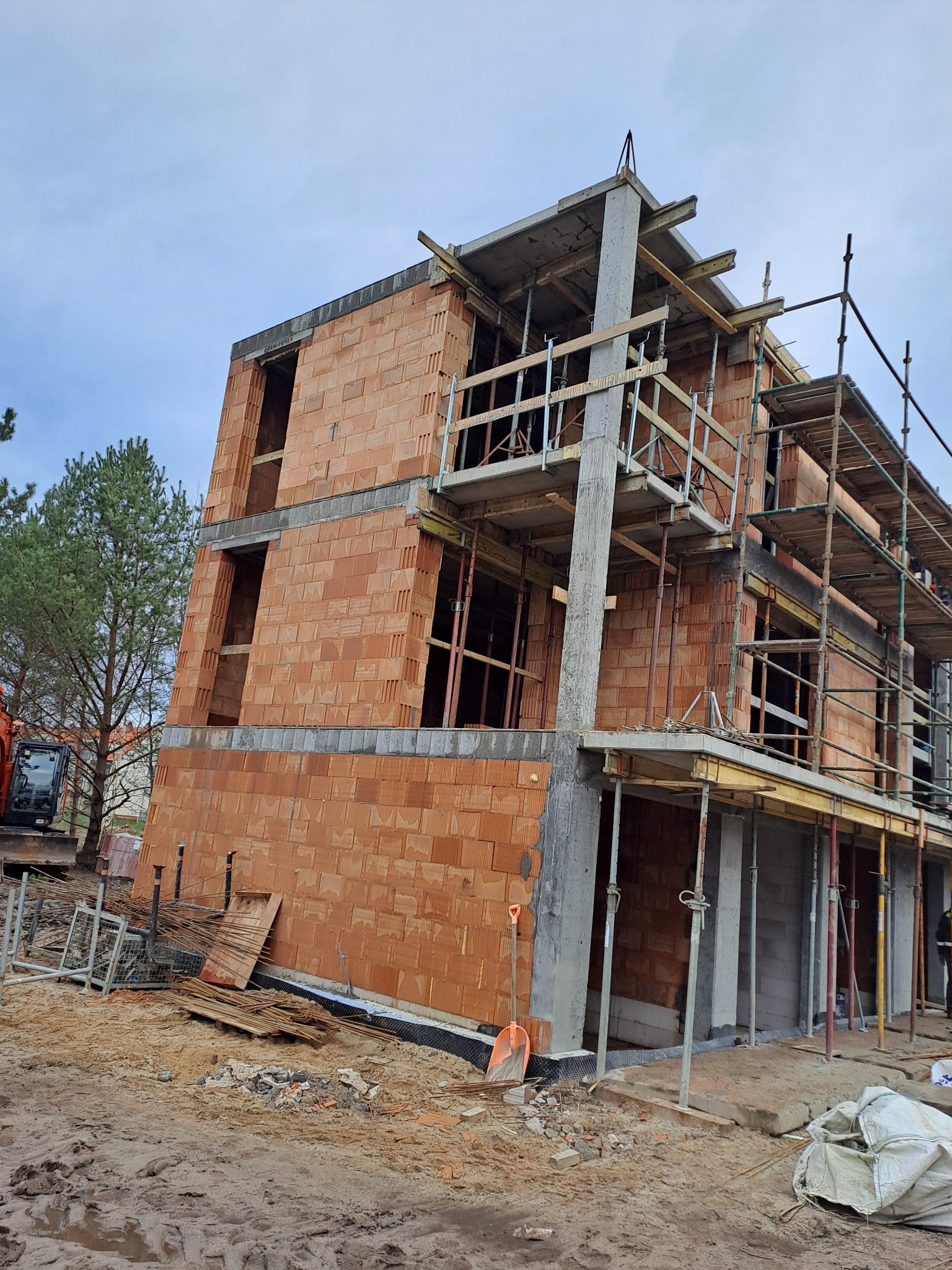 Dwukondygnacyjny budynek w trakcie budowy, wzniesiony z czerwonej cegły ceramicznej, z widocznymi betonowymi słupami i rusztowaniami, na tle pochmurnego nieba i drzew