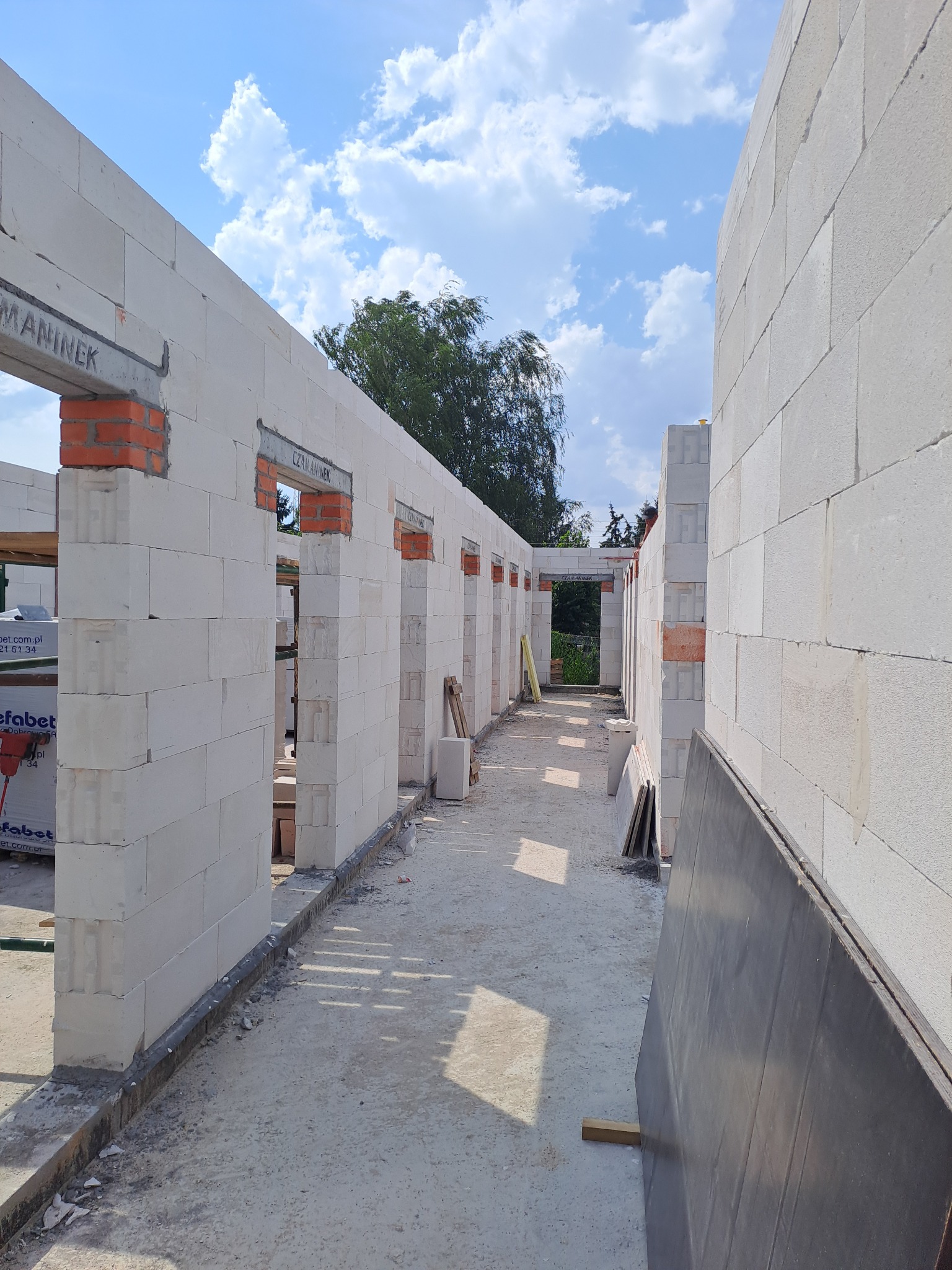 Wnętrze domu w trakcie budowy, widoczne ściany z białych bloczków z pustymi otworami okiennymi z czerwonej cegły, na podłożu widoczny gruz i płyty budowlane, w tle zieleń drzew.