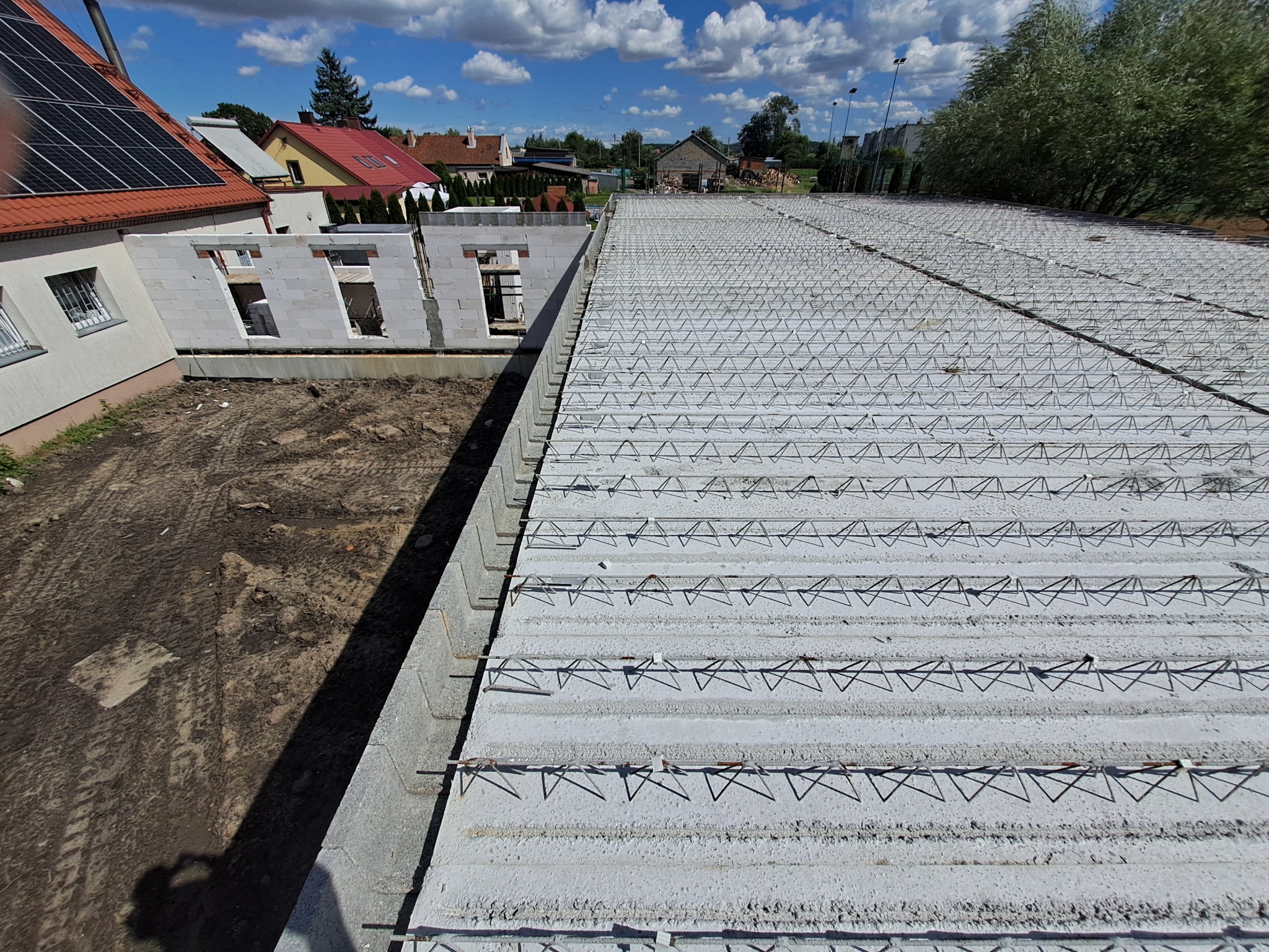 Wysoki kąt widzenia na budowę domu z białych bloczków, widoczne zbrojenie stropu, w tle zabudowa jednorodzinna i błękitne niebo z chmurami.