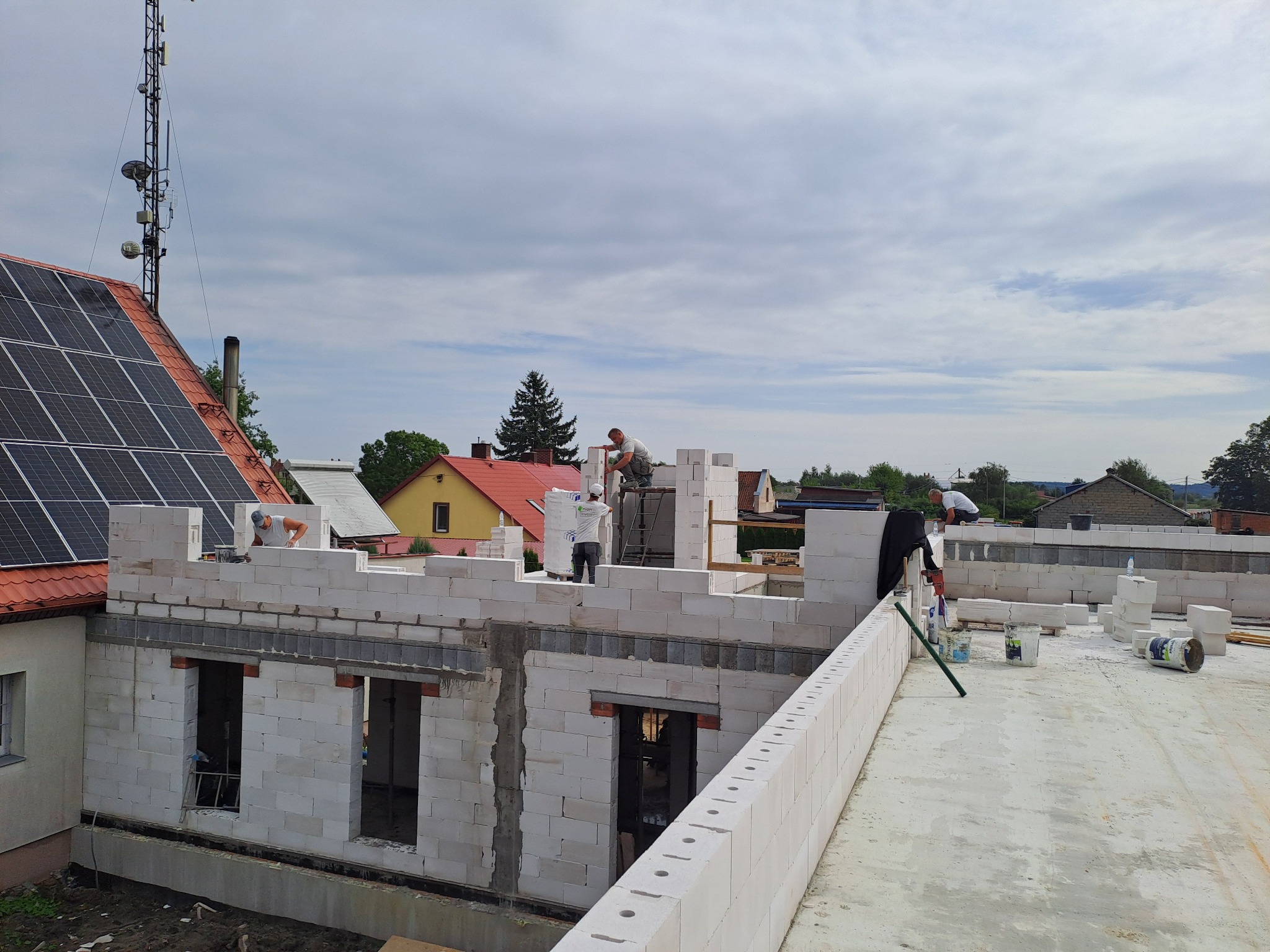 Wznoszenie ścian parterowego budynku z białych bloczków, widoczni pracownicy budowlani, elementy szalunków okiennych, w tle domy mieszkalne i pochmurne niebo.