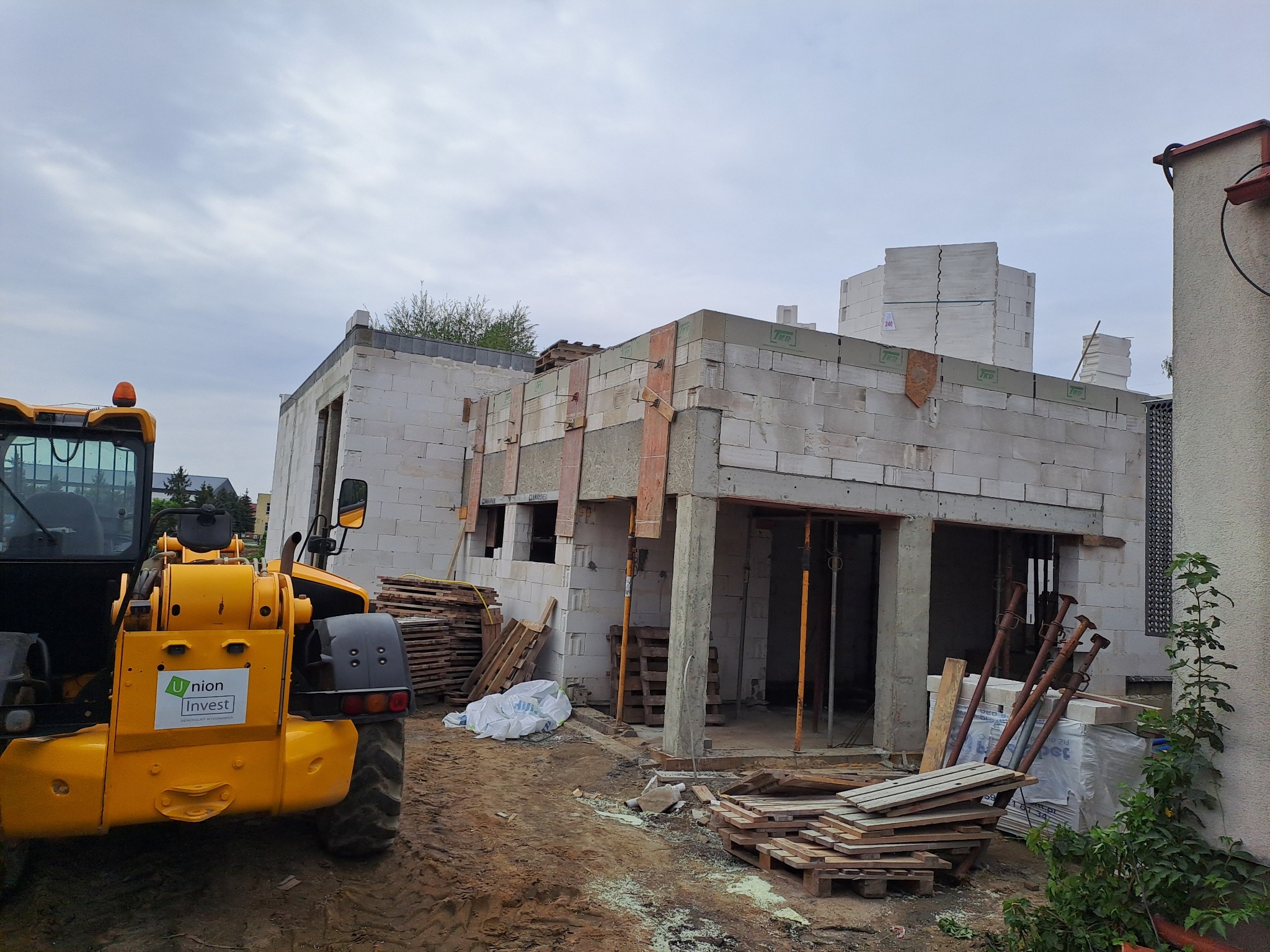 Wznoszenie ścian parterowego domu z bloczków betonu komórkowego, widoczny żółty teleskopowy podnośnik z logo firmy budowlanej, drewniane palety i podpory szalunkowe przy budynku.