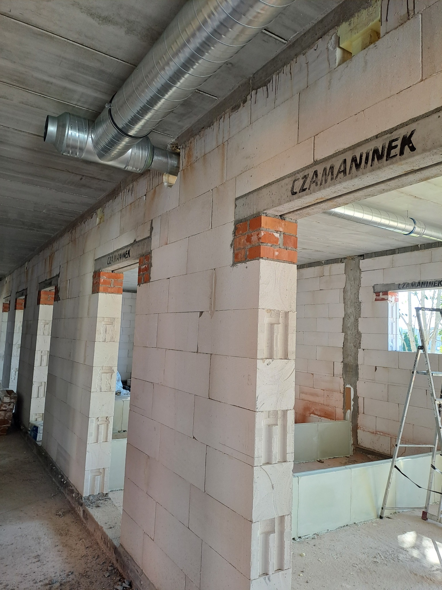 Wnętrze surowego domu w trakcie budowy, widoczne białe ściany z betonu komórkowego, otwory drzwiowe wzmocnione czerwoną cegłą, instalacja wentylacyjna pod sufitem, pianka izolacyjna przy podłodze...