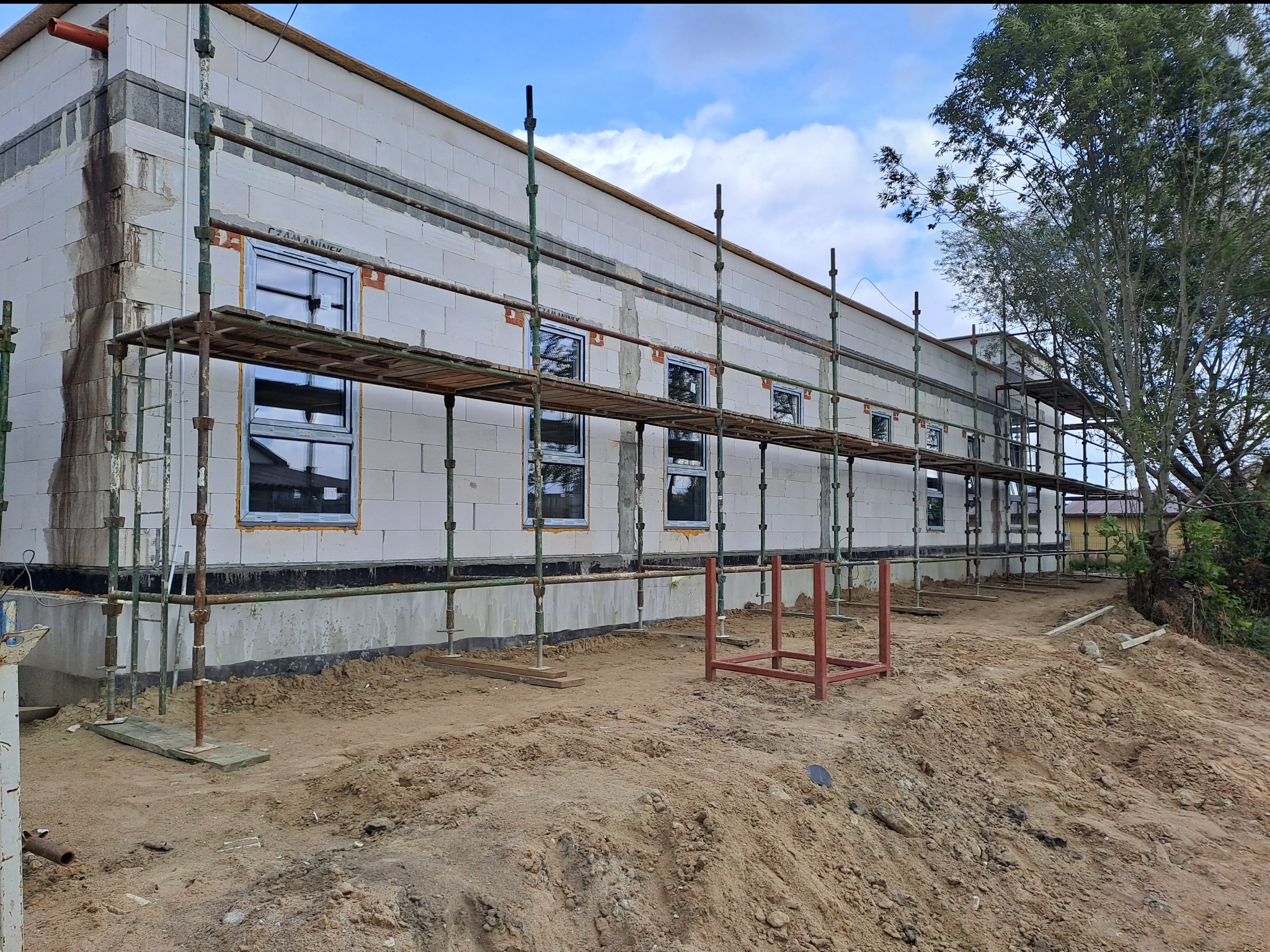 Surowy stan budynku z białego bloczka, widoczne rusztowania, wstawione okna, teren wokół wyrównany, w tle drzewo i fragment nieba z chmurami.