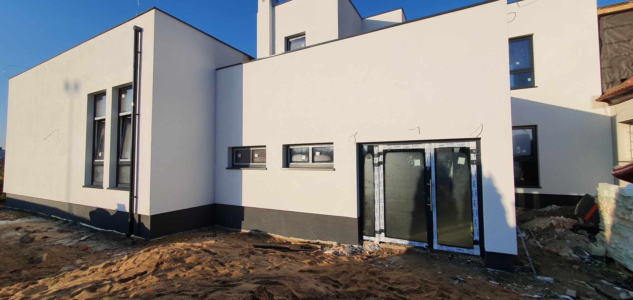 Nowoczesny, biały budynek z płaskim dachem w trakcie budowy, widoczne okna z czarnymi ramami, niezabudowane otoczenie z piaskiem i gruzem.