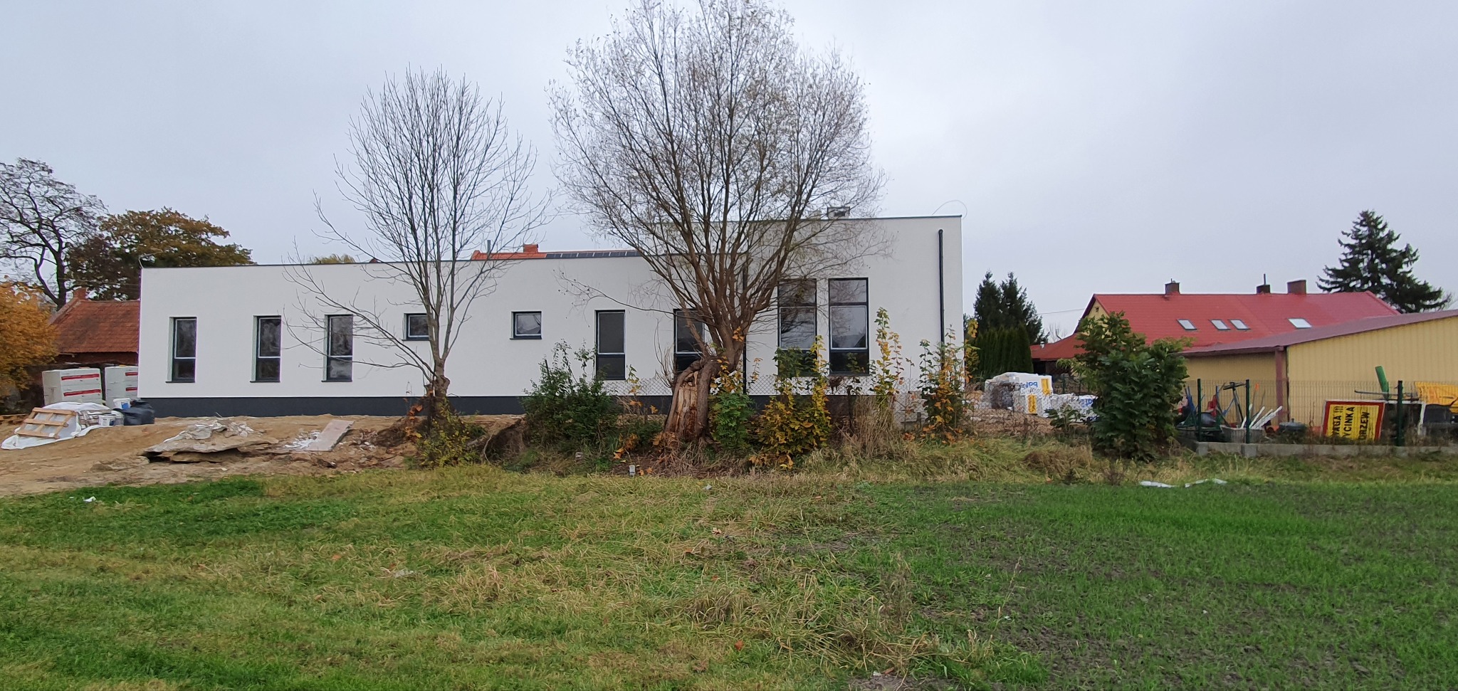 Nowoczesny dom jednorodzinny w trakcie budowy, z białymi ścianami i dużymi oknami, otoczony trawnikiem i drzewami bez liści, z widocznymi materiałami budowlanymi na działce.