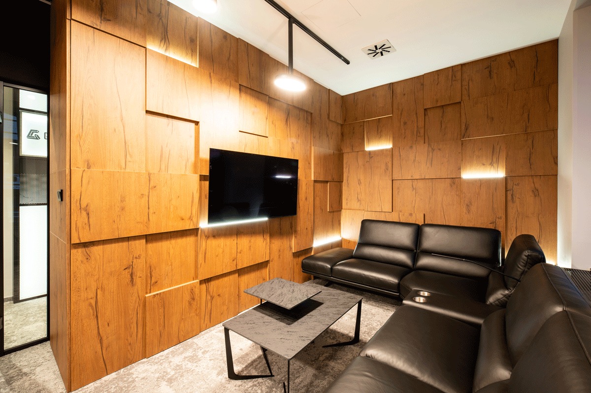 Nowoczesna aranżacja biura z drewnianą ścianą z podświetleniem LED, czarnym telewizorem, skórzaną sofą i stolikiem kawowym na dywanie w neutralnych kolorach.