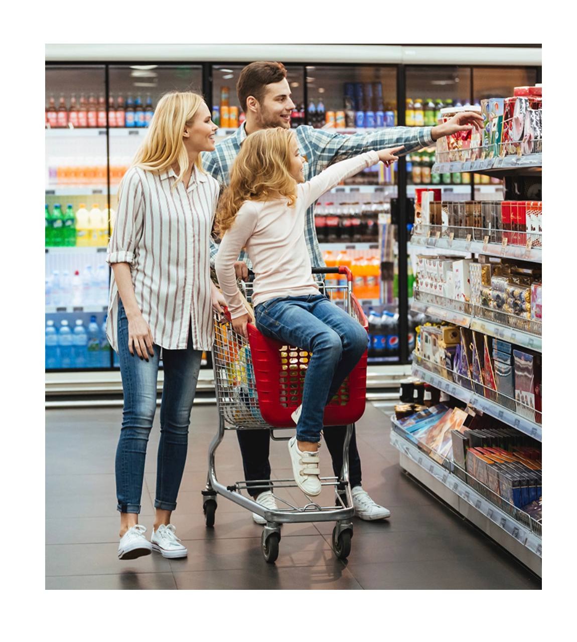 Rodzina z dzieckiem siedzącym w czerwonym wózku na zakupy w supermarkecie, mężczyzna wskazuje na półkę z produktami.