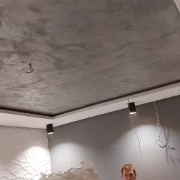 Sufit z dekoracyjną szarą strukturą przypominającą beton, czarne reflektory punktowe, biała ściana z trójwymiarowym wzorem fal oraz fragment osoby w prawym dolnym rogu.