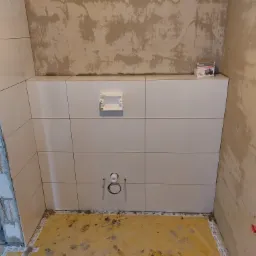 Białe płytki ułożone na ścianie łazienki, widoczne wystające rury instalacji wodnej i przygotowane otwory pod stelaż WC, surowy stan ścian i podłogi w trakcie prac wykończeniowych.