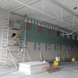 Montaż płyt karton gips na ścianie w dużym pomieszczeniu, widoczny pracownik na rusztowaniu, stos płyt, izolacja i nagrzewnice.