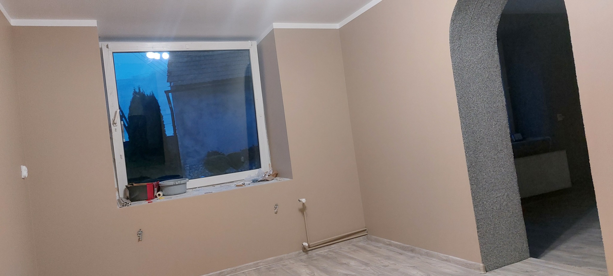 Beżowe ściany świeżo po malowaniu, widoczne narzędzia malarskie na parapecie okna i szary kamienny łuk drzwiowy.