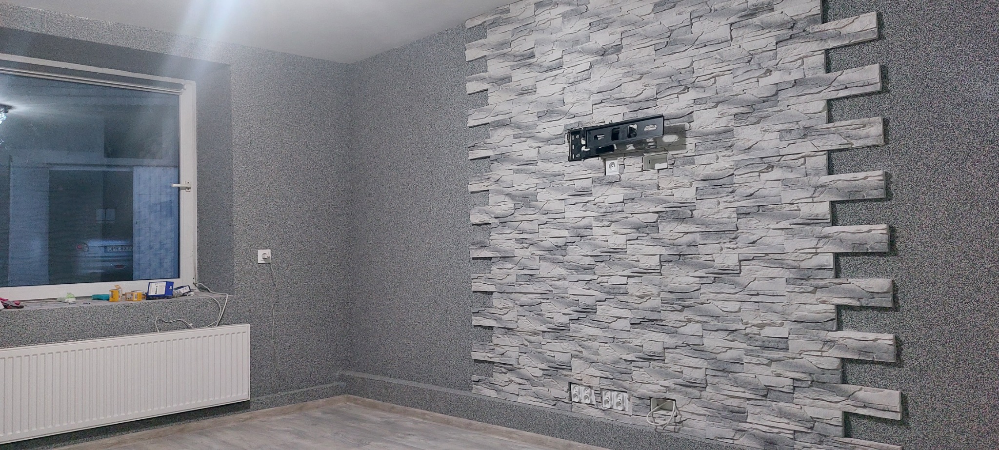 Fragment pokoju z dekoracyjną ścianą z paneli imitujących kamień, na której zamontowany jest uchwyt do telewizora. Ściany pokryte szarym, ziarnistym tynkiem.