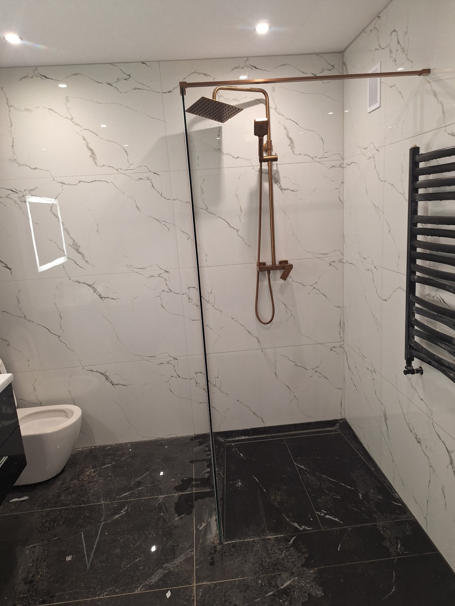 Wykończona łazienka w Prudniku z kabiną prysznicową typu walk-in, armaturą w kolorze miedzi i grzejnikiem drabinkowym na tle marmurowych płytek ściennych.
