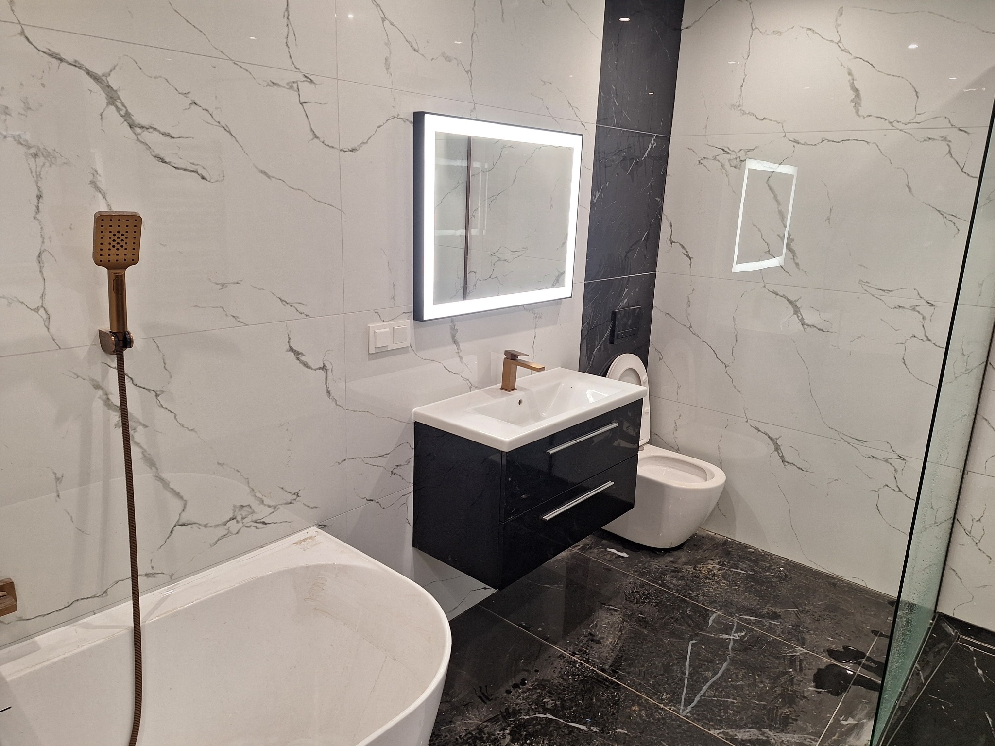 Nowoczesna łazienka z marmurowymi płytkami, podświetlanym lustrem, czarną szafką pod umywalką i złotą armaturą.