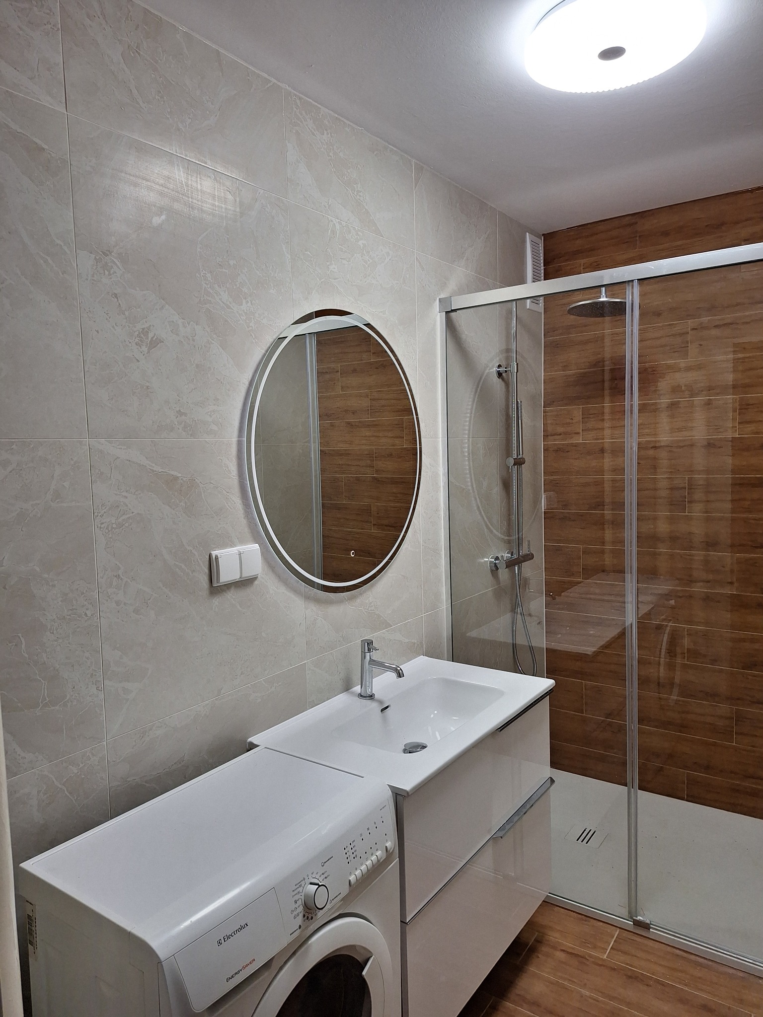 Jasna łazienka po remoncie: beżowe płytki imitujące marmur, okrągłe lustro z podświetleniem, kabina prysznicowa z brązowymi płytkami i pralka obok umywalki.