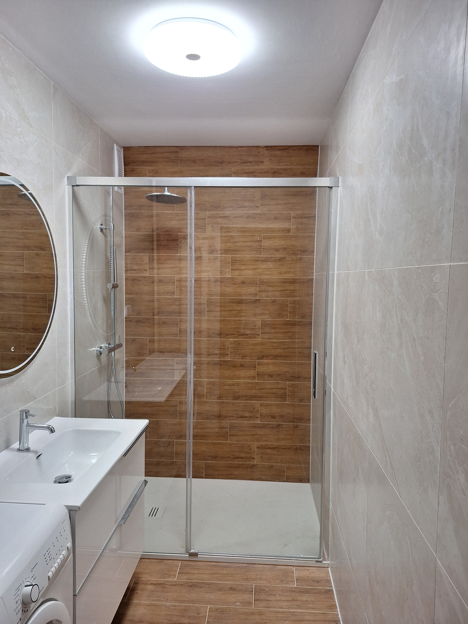 Nowoczesna łazienka po remoncie: kabina prysznicowa z drewnopodobnymi płytkami, umywalka z szafką i okrągłe lustro. Jasne płytki na ścianach i podłodze.