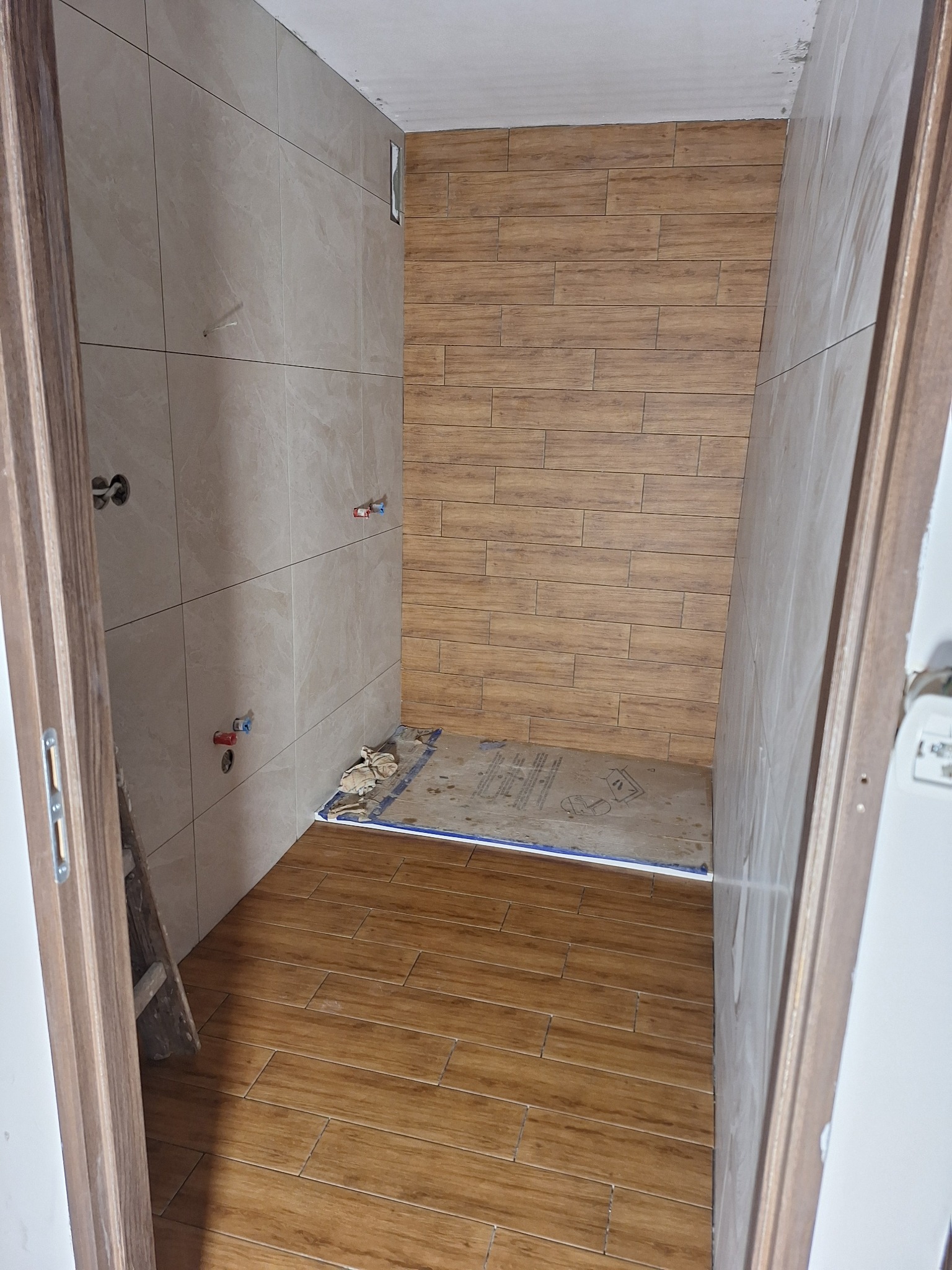 Wykończona łazienka z beżowymi i brązowymi płytkami imitującymi drewno na ścianach i podłodze. Widoczne przyłącza wodne i fragment drabiny. Ujęcie zza otwartych drzwi.