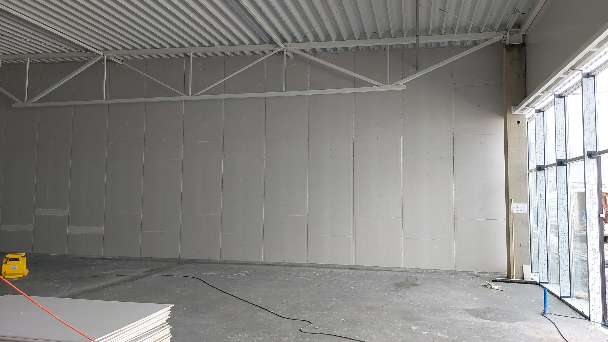 Wnętrze w trakcie prac: ściana wyłożona płytami kartonowo-gipsowymi, widoczne łączenia płyt wymagające szpachlowania, surowy betonowy strop z widoczną konstrukcją nośną, podłoga betonowa, okna...