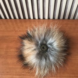 Usługi krawiecko-kuśnierskie - Puszysty pompon z futra w odcieniach brązu, beżu i szarości, przymocowany zatrzaskiem do drewnianej powierzchni na tle pionowych pasków.