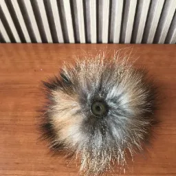 Puszysty pompon z futra w odcieniach brązu, beżu i szarości, przymocowany zatrzaskiem do drewnianej powierzchni na tle pionowych pasków.