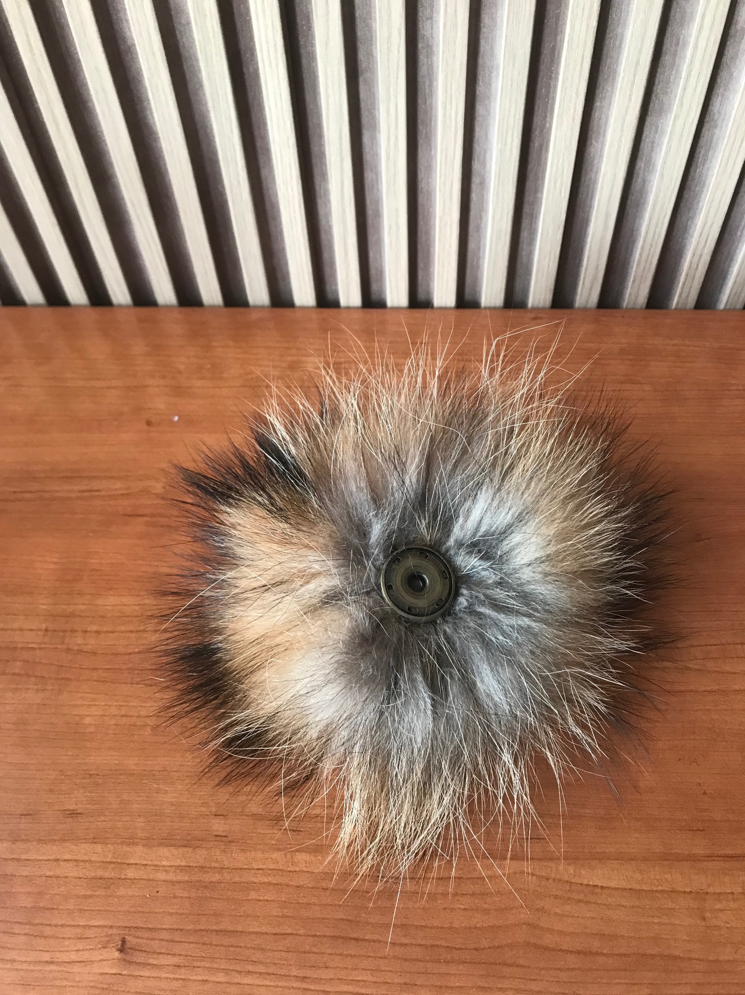 Puszysty pompon z futra w odcieniach brązu, beżu i szarości, przymocowany zatrzaskiem do drewnianej powierzchni na tle pionowych pasków.