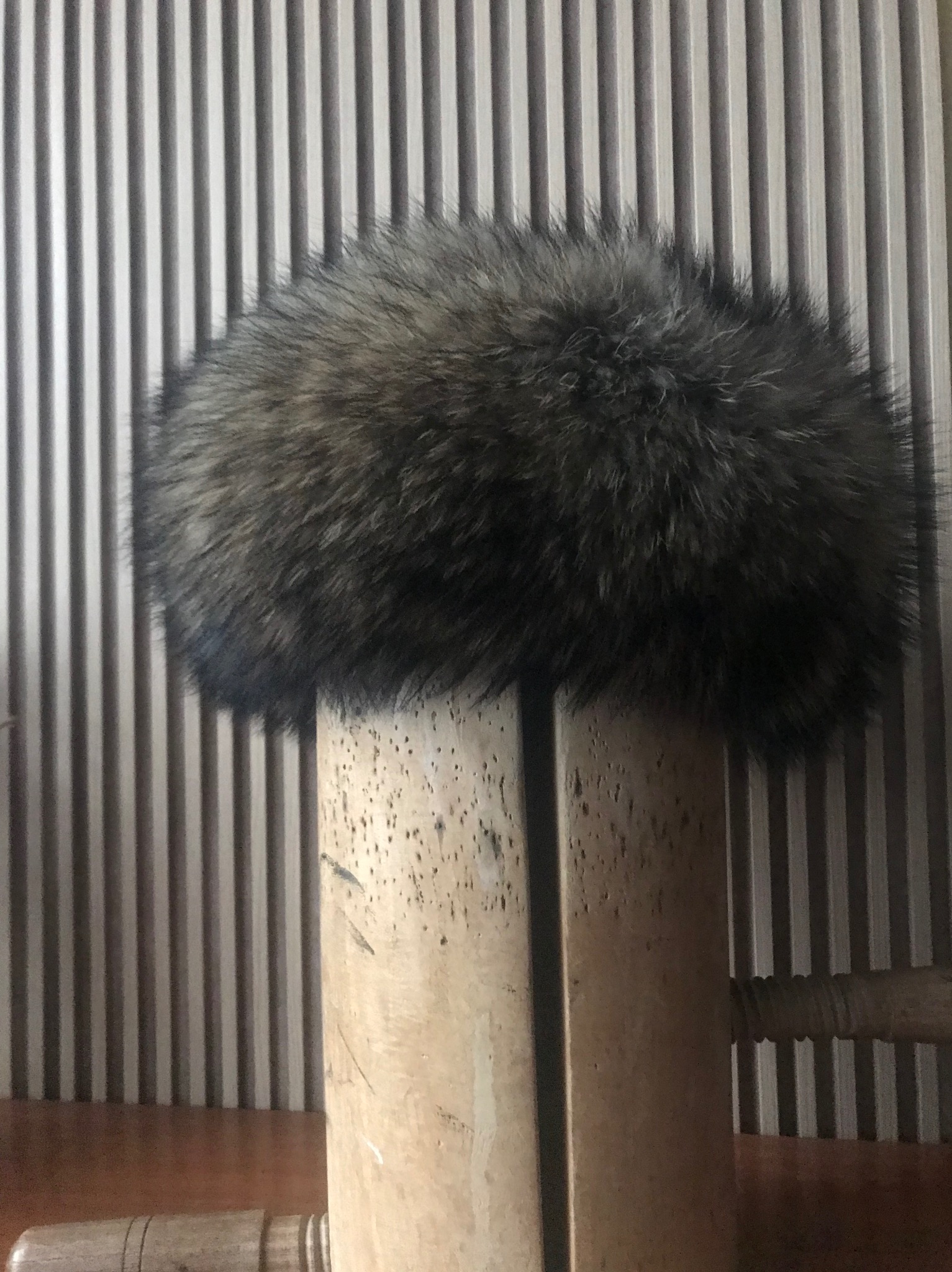 Puszysta, okrągła czapka z futra w odcieniach brązu i szarości, ustawiona na drewnianej formie, na tle ściany z pionowymi paskami.