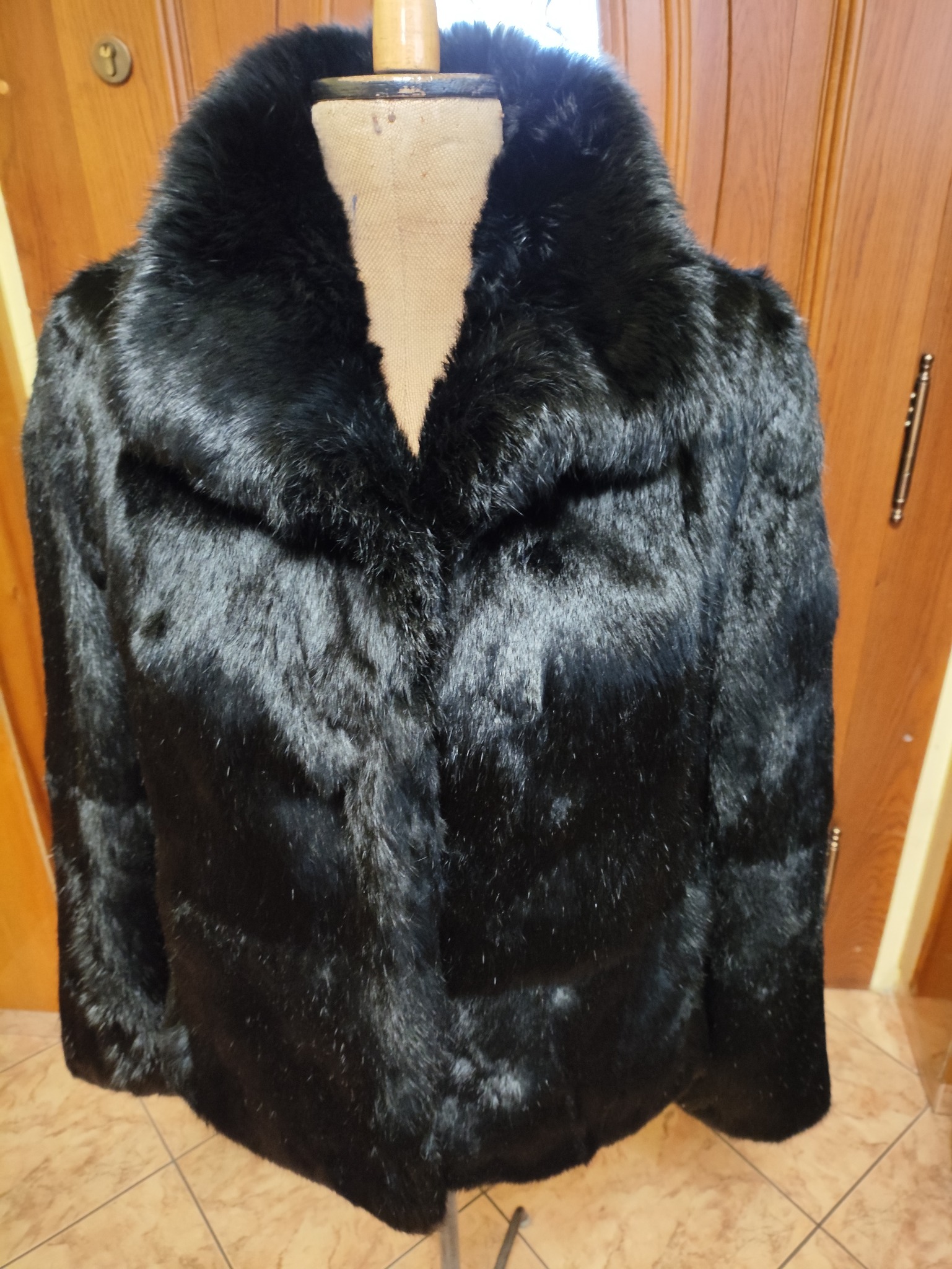 Czarny, futrzany płaszcz z szerokim kołnierzem, prezentowany na manekinie krawieckim na tle drewnianych drzwi i podłogi z płytek. Widoczne detale futra i krój płaszcza.