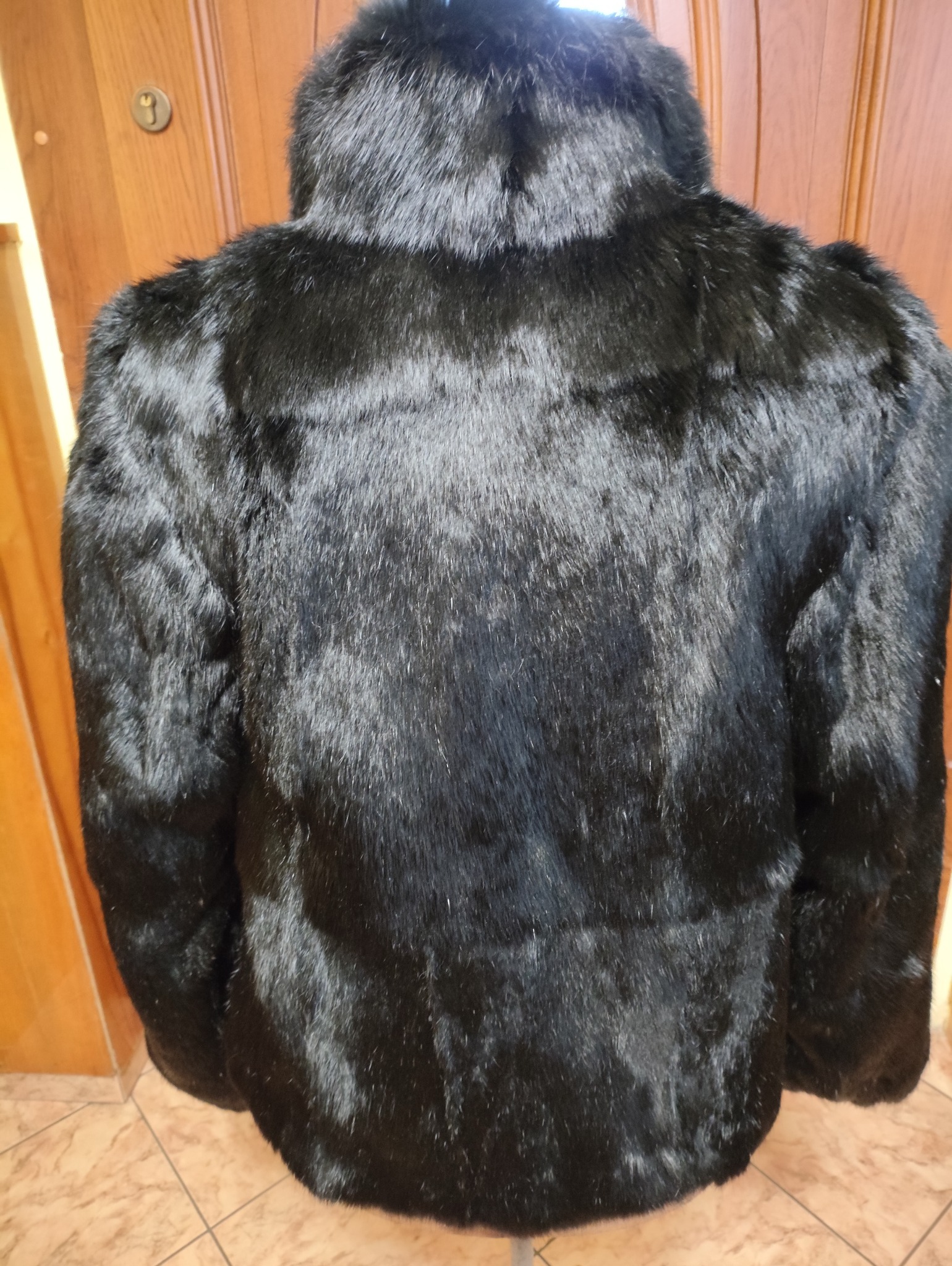 Czarna, futrzana kurtka z wysokim kołnierzem, widok z tyłu. Widoczna faktura futra i kształt kurtki. Tło stanowi drewniana ściana i fragment podłogi z płytek.