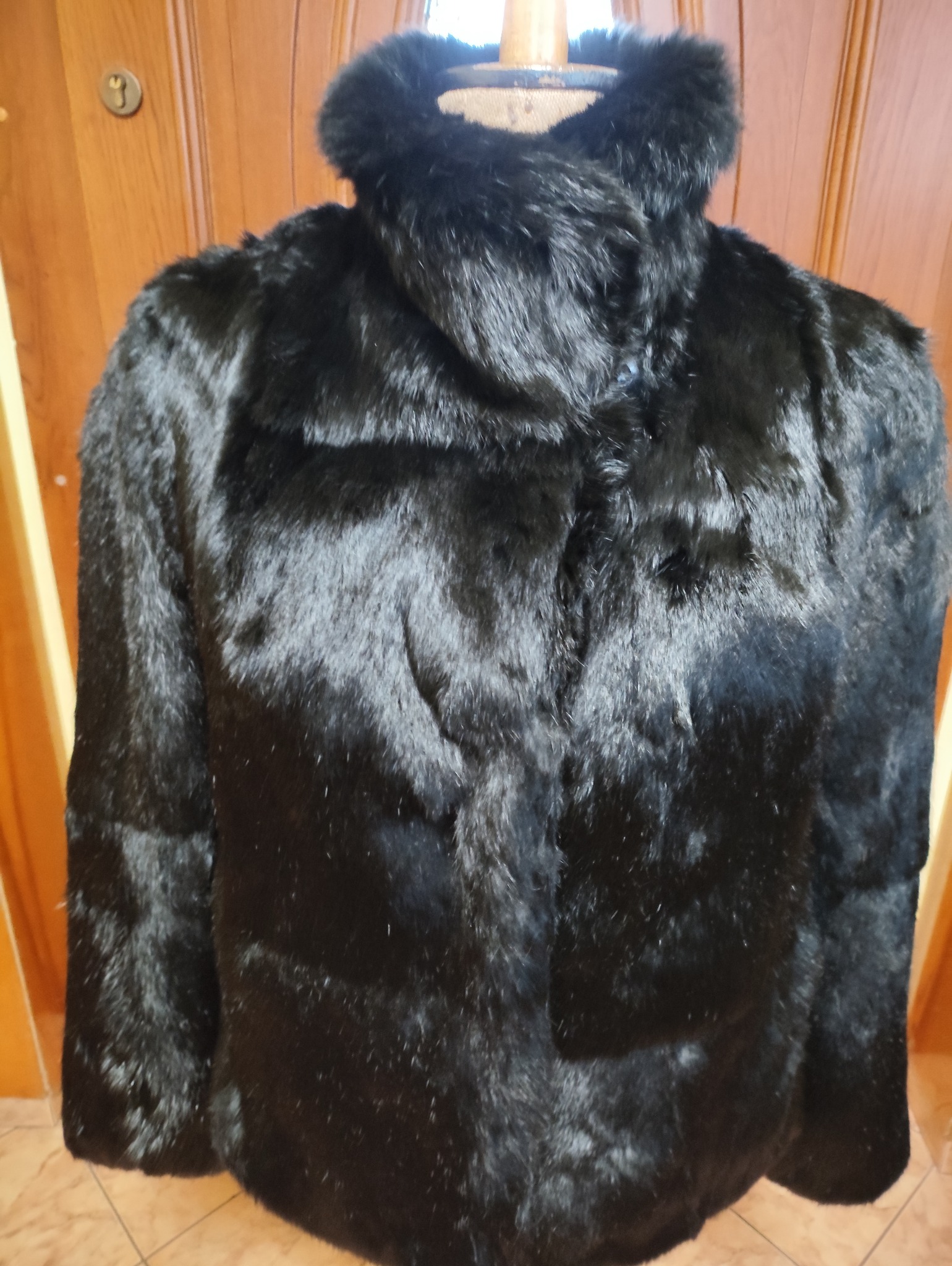 Czarny, futrzany płaszcz z wysokim kołnierzem, prezentowany na manekinie na tle drewnianych drzwi. Widoczne detale faktury futra i zapięcie na guziki.