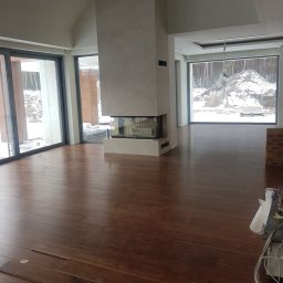 PAECH NEW HOME - Wnętrze domu w trakcie układania parkietu z drewna egzotycznego, widoczny kominek obłożony kamieniem, duże okna z widokiem na zimowy krajobraz.
