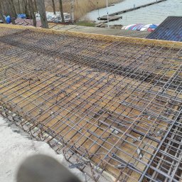 PAECH NEW HOME - Zbrojenie stropu z widoczną siatką z prętów stalowych, szalunkiem drewnianym i elementami dystansowymi, w tle jezioro i drzewa.