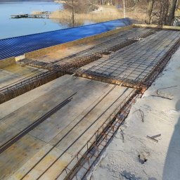 MARIA PAECH NEW HOME - Przygotowanie szalunku i zbrojenia pod wylewkę betonową na tle jeziora i lasu, widoczne deski szalunkowe, siatka zbrojeniowa i elementy stalowe.