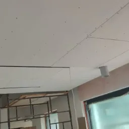 Montaż sufitu podwieszanego z płyt kartonowo-gipsowych, widoczne szpachlowane łączenia, rury instalacyjne i rusztowanie w tle.