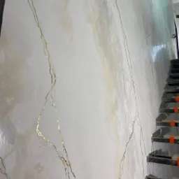 Ściana wewnętrzna z dekoracyjną gładzią imitującą marmur, z wyraźnymi złotymi żyłkami i delikatnymi beżowymi przebarwieniami, widoczne schody w tle.