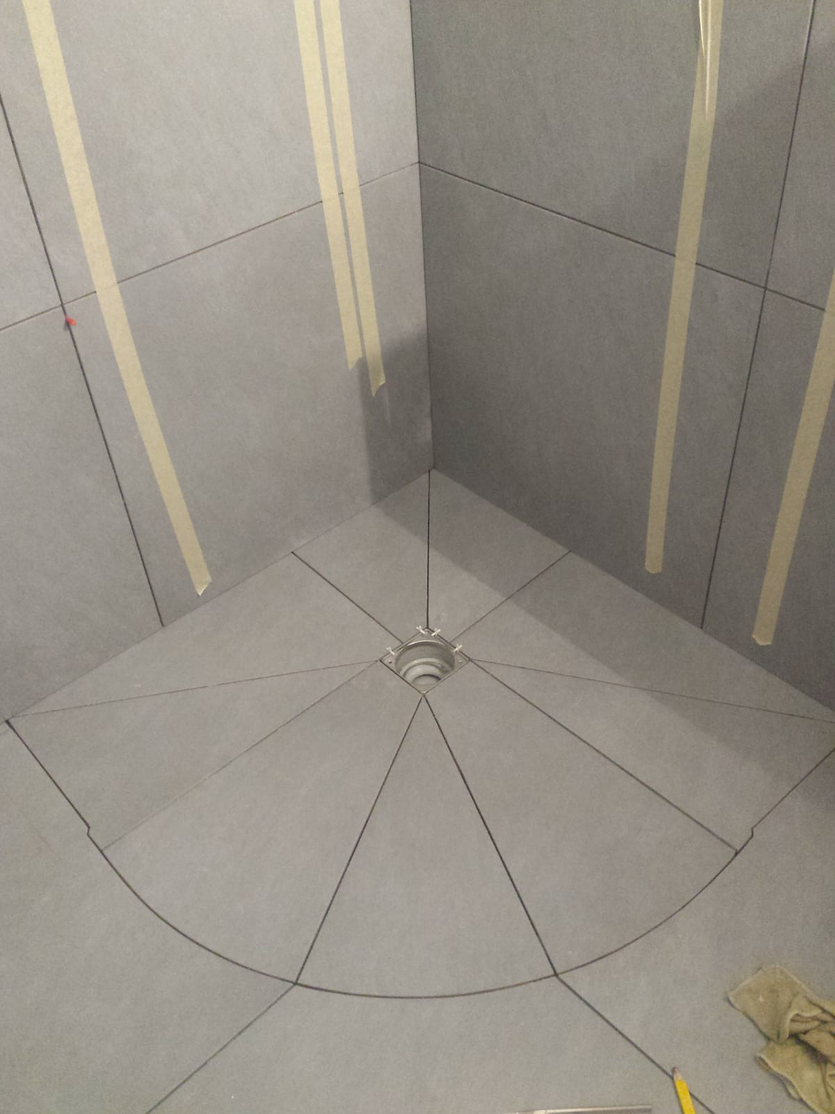Płytki ceramiczne ułożone w kabinie prysznicowej, z centralnym odpływem i zaznaczonymi liniami cięcia, zabezpieczone taśmą malarską na ścianach.