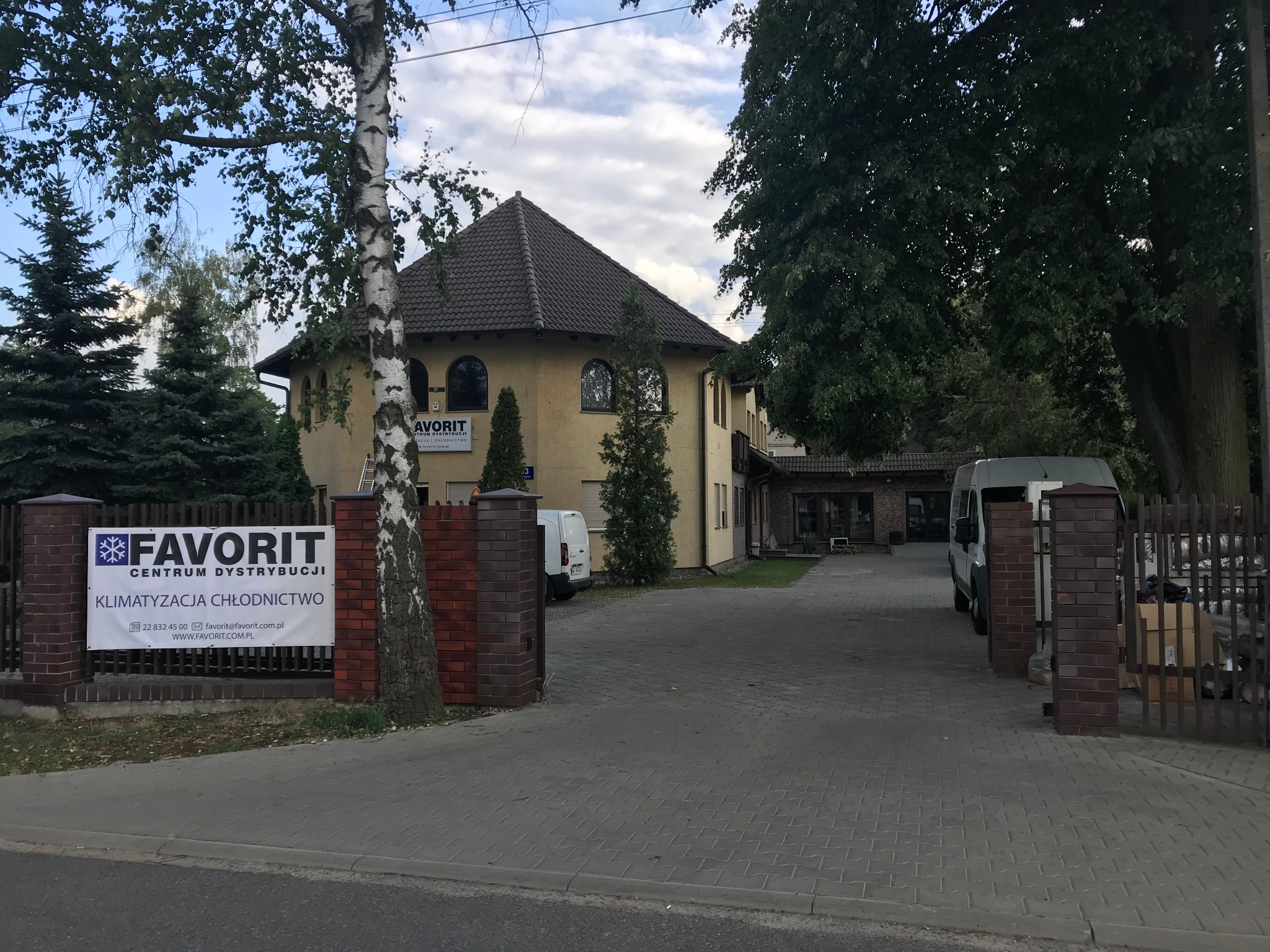 Budynek firmy Favorit Centrum Dystrybucji Klimatyzacja Chłodnictwo z banerem reklamowym na ogrodzeniu, biała drabina oparta o ścianę budynku, dwa busy na podjeździe.