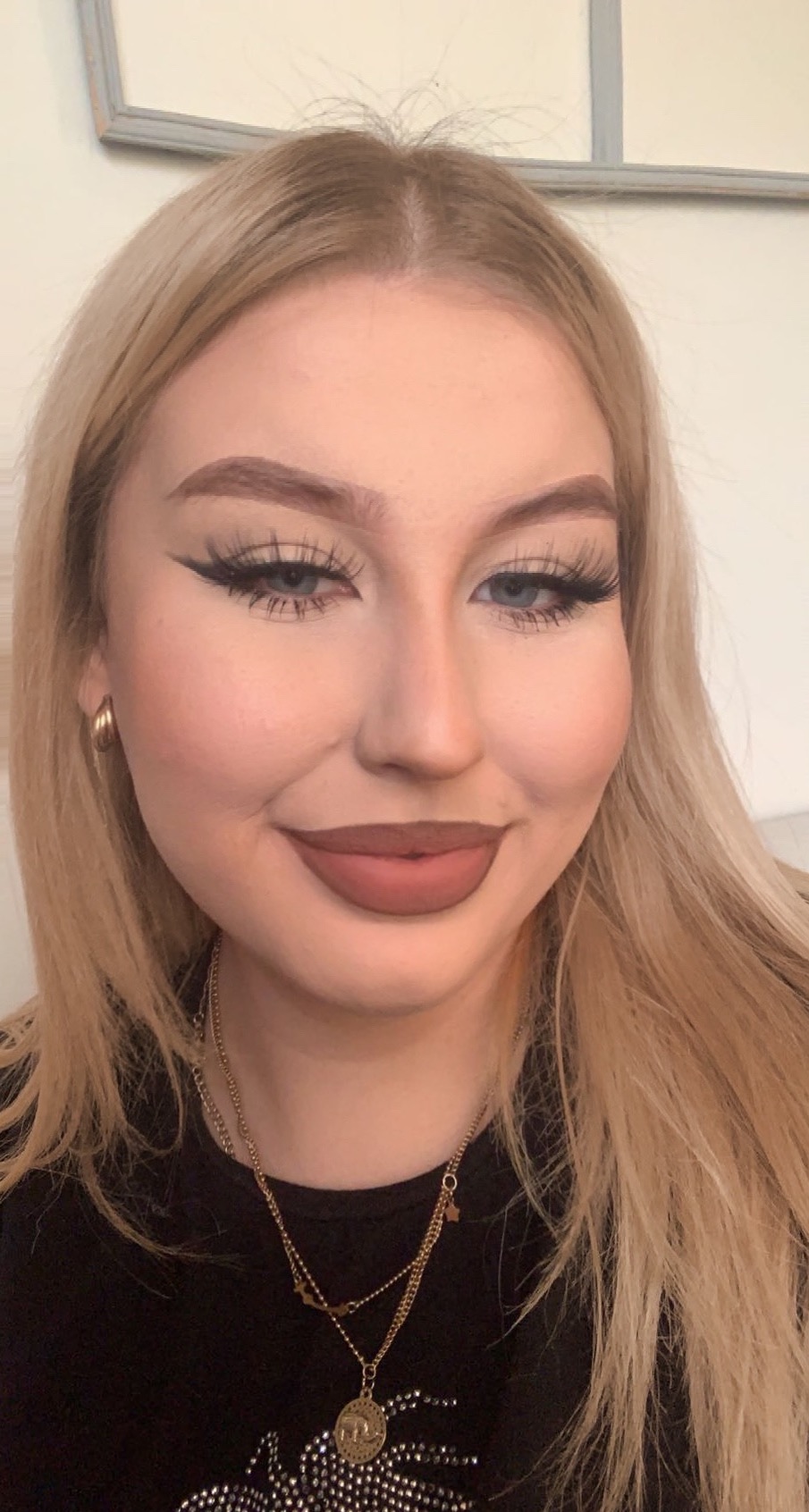 Portret młodej kobiety z blond włosami, z wyrazistym makijażem oczu i ust, noszącej złote kolczyki i naszyjniki, uśmiechającej się delikatnie.