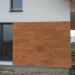 Fragment elewacji domu jednorodzinnego w Nowym Tomyślu z drewnianą okładziną, widok na okno i fragment dachu w trakcie wykończenia.