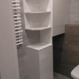 Biała, narożna szafka łazienkowa z półkami, umieszczona obok grzejnika drabinkowego i podwieszanej szafki, na tle szarych i brązowych płytek.