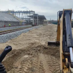 Widok z kabiny koparki w Szczecinie na teren budowy z widoczną infrastrukturą przemysłową i fragmentem torów kolejowych. Ślady opon na piasku sugerują prace ziemne.